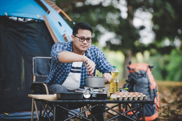 Retrato de gafas de hombre viajero asiático freír un sabroso huevo frito en una sartén caliente en el camping Cocina al aire libre concepto de estilo de vida de camping de viaje