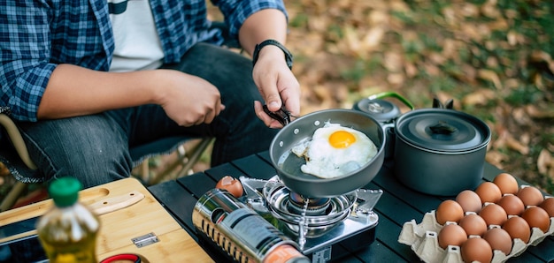 Retrato de gafas de hombre viajero asiático freír un sabroso huevo frito en una sartén caliente en el camping Cocina al aire libre concepto de estilo de vida de camping de viaje