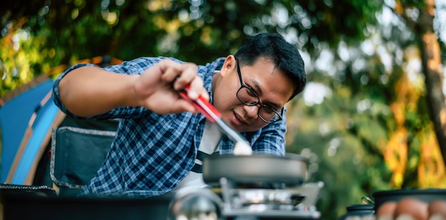 Retrato de gafas de hombre viajero asiático freír un sabroso huevo frito en una sartén caliente en el camping Cocina al aire libre concepto de estilo de vida de camping de viaje