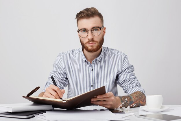 Retrato del gerente masculino seguro con tatuajes, escribe en el plan diario para la próxima semana, bebe café