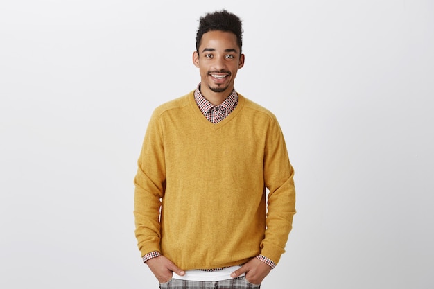 Retrato de guapo afroamericano con peinado afro en elegante jersey amarillo tomados de la mano en los bolsillos, sonriendo casualmente