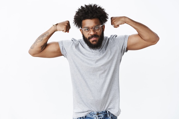 Foto gratuita retrato de guapo chico afroamericano autosatisfecho trabajando fuerte y masculino mostrando bíceps con las manos levantadas mirando serio y desafiante al frente como invitando al gimnasio