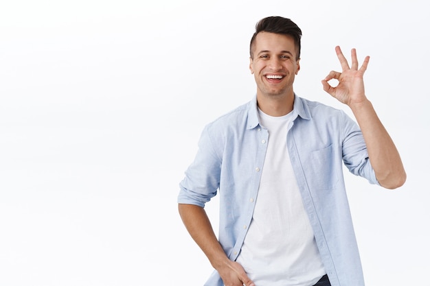 Retrato de un guapo chico de familia satisfecho con camisa sobre camiseta, muestra una confirmación aceptable o un gesto de seguridad, sonriendo complacido, recomienda un producto de buena calidad, da permiso, garantiza la calidad