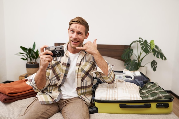 Foto gratuita retrato de un guapo chico rubio con una cámara en la mano mostrando el gesto de una llamada telefónica y sonriendo sentado