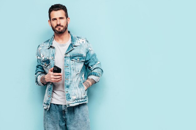Retrato de guapo confiado elegante hipster lambersexual modelo Hombre sexy vestido con chaqueta de jeans Hombre de moda aislado en la pared azul en el estudio Sosteniendo el teléfono inteligente