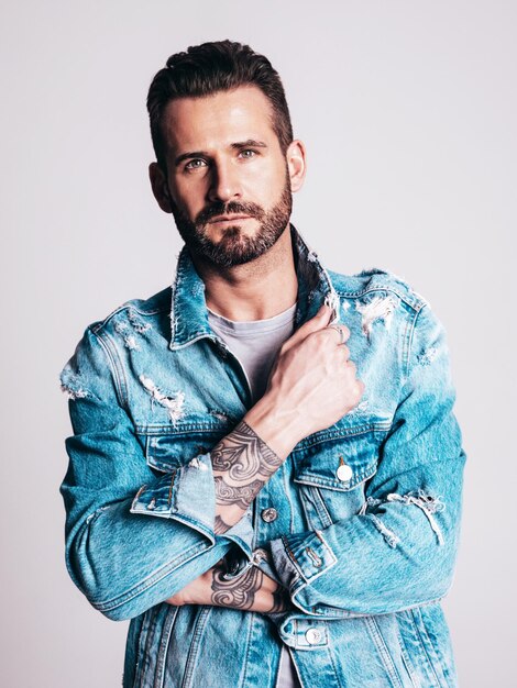 Retrato de guapo confiado elegante hipster lambersexual modelo Sexy hombre vestido con chaqueta de jeans Moda hombre aislado sobre fondo gris en estudio