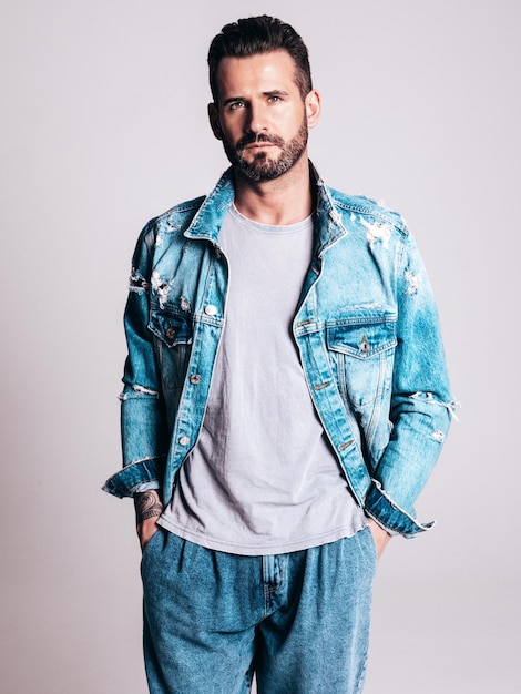 Foto gratuita retrato de guapo confiado elegante hipster lambersexual modelo sexy hombre vestido con chaqueta de jeans moda hombre aislado sobre fondo gris en estudio