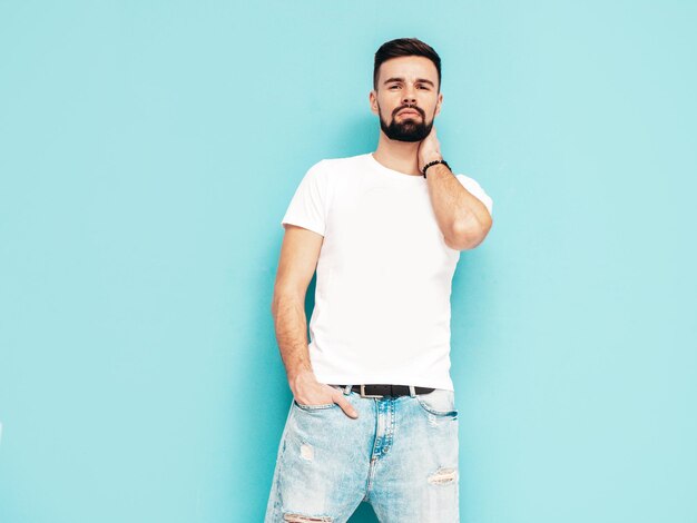 Retrato de guapo confiado con estilo hipster lambersexual modelMan vestido con camiseta blanca y jeans Moda hombre aislado en la pared azul en el estudio