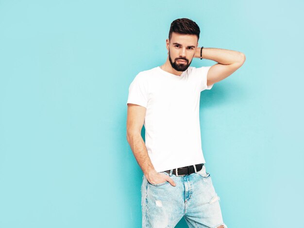 Retrato de guapo confiado con estilo hipster lambersexual modelMan vestido con camiseta blanca y jeans Moda hombre aislado en la pared azul en el estudio