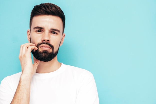 Retrato de guapo confiado con estilo hipster lambersexual modelMan vestido con camiseta blanca y jeans Moda hombre aislado en la pared azul en el estudio