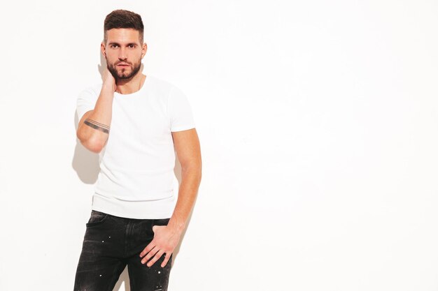 Retrato de guapo confiado con estilo hipster lambersexual modelMan vestido con camiseta blanca y jeans Moda masculina posando cerca de la pared en el estudio aislado