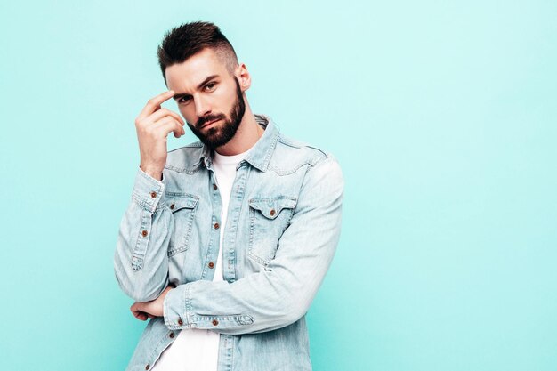 Retrato de guapo confiado con estilo hipster lambersexual modelMan vestido con chaqueta y jeans Fashion male posando cerca de la pared azul en el estudio