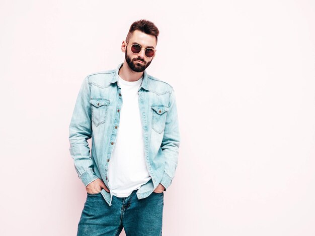 Retrato de guapo confiado con estilo hipster lambersexual modelMan vestido con chaqueta y jeans Hombre de moda posando en el estudio con gafas de sol Aislado en blanco