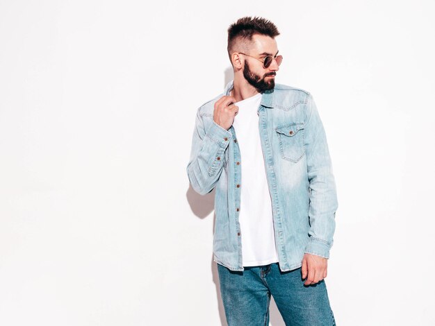 Retrato de guapo confiado con estilo hipster lambersexual modelMan vestido con chaqueta y jeans Hombre de moda posando en el estudio con gafas de sol Aislado en blanco