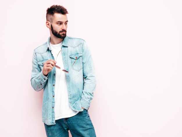 Retrato de guapo confiado con estilo hipster lambersexual modelMan vestido con chaqueta y jeans Hombre de moda posando en el estudio con gafas de sol Aislado en rosa claro