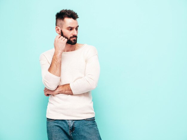 Retrato de guapo confiado con estilo hipster lambersexual modelMan vestido con suéter blanco y jeans Moda hombre aislado en la pared azul en el estudio