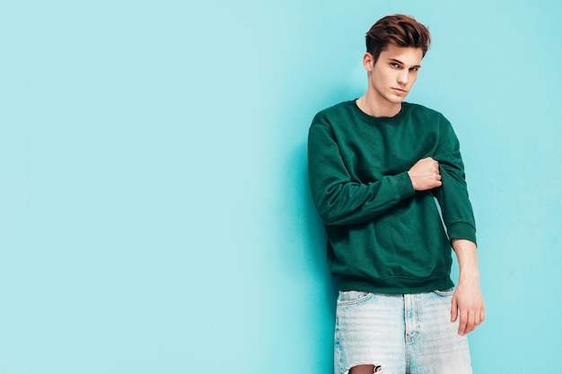 Foto gratuita retrato de guapo confiado con estilo hipster lambersexual modelman vestido con suéter verde y jeans hombre de moda posando en el estudio cerca de la pared azul