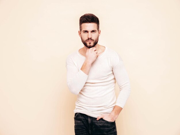 Retrato de guapo confiado con estilo hipster lambersexual modelo Hombre vestido con suéter blanco y jeans Moda hombre posando junto a la pared en el estudio