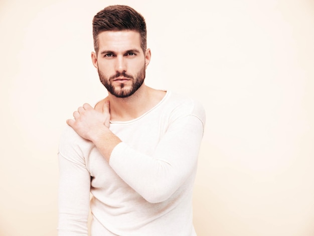 Retrato de guapo confiado con estilo hipster lambersexual modelo Hombre vestido con suéter blanco y jeans Moda hombre posando junto a la pared en el estudio