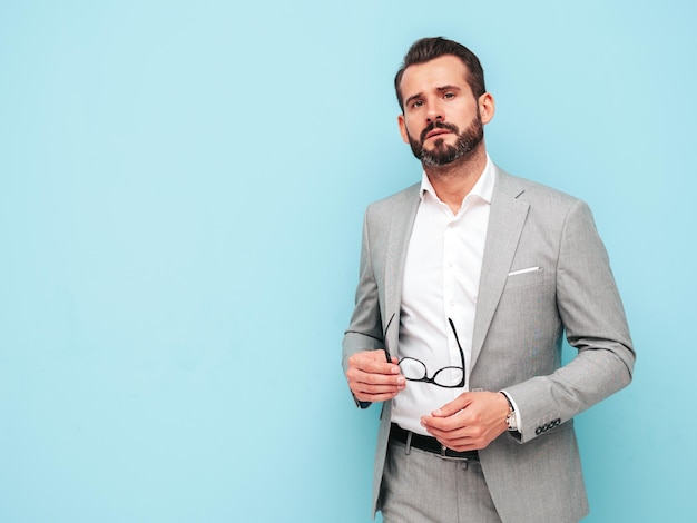 Retrato de guapo confiado con estilo hipster lambersexual modelSexy hombre moderno vestido con traje elegante Hombre de moda posando en el estudio cerca de la pared azul