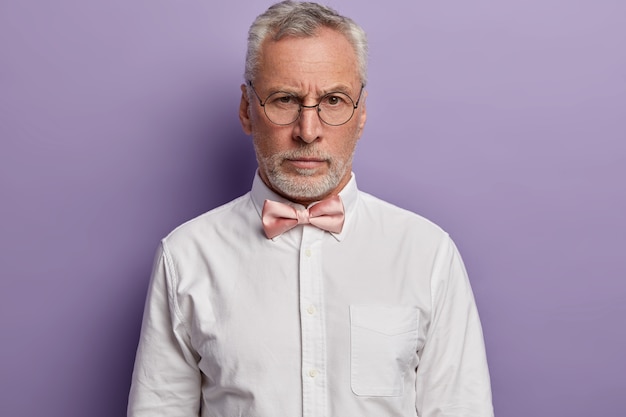 Retrato de guapo hombre europeo de pelo gris mira con expresión estricta a través de gafas redondas, viste camisa blanca formal y pajarita