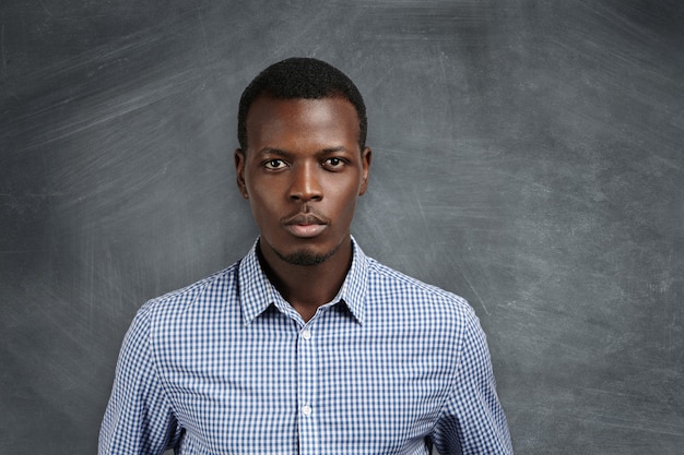 Retrato de guapo joven profesor de escuela africana con camisa a cuadros preparándose para la lección, tomando una decisión, mirando con expresión seria y segura, de pie en la pizarra en blanco