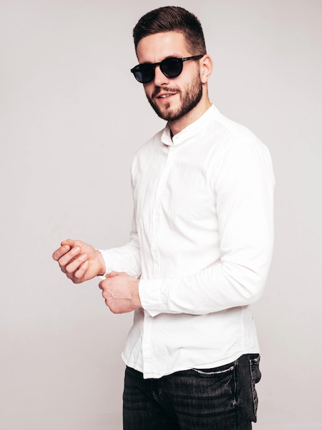 Retrato de guapo modelo confiado Hombre elegante sexy vestido con camisa blanca y jeans Hombre hipster de moda posando en estudio sobre fondo gris En gafas de sol Aislado