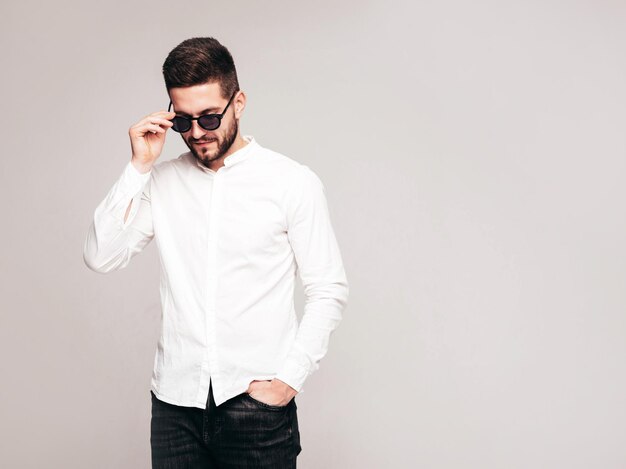 Retrato de guapo modelo confiado Hombre elegante sexy vestido con camisa blanca y jeans Hombre hipster de moda posando en estudio sobre fondo gris En gafas de sol Aislado