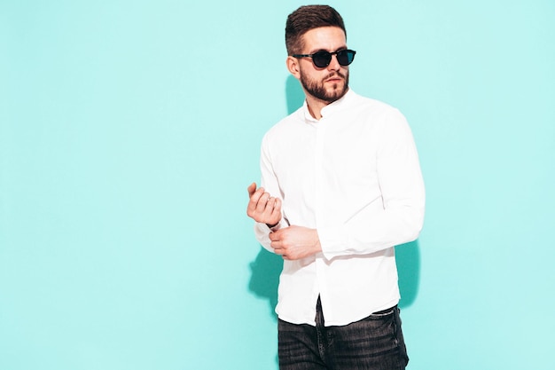 Retrato de guapo modelo confiado Hombre elegante sexy vestido con camisa blanca y jeans Hombre hipster de moda posando junto a la pared azul en el estudio Aislado en gafas de sol