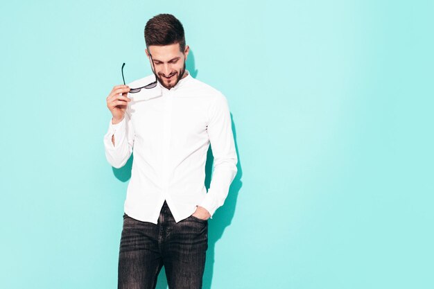 Retrato de guapo modelo confiado Hombre elegante sexy vestido con camisa blanca y jeans Hombre hipster de moda posando junto a la pared azul en el estudio Aislado en gafas de sol