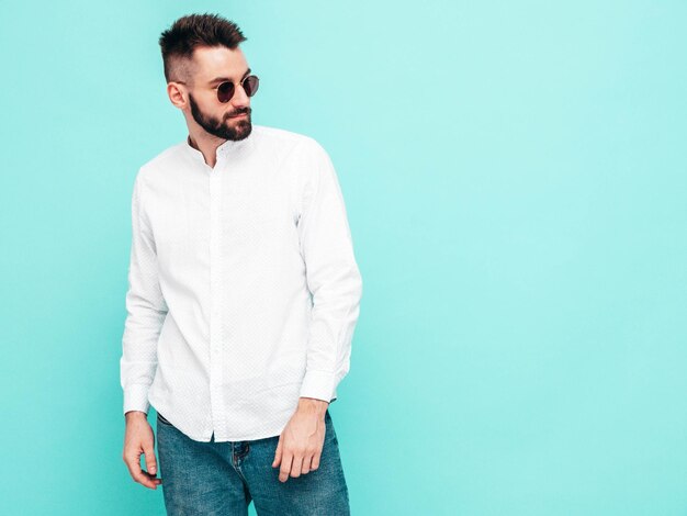 Retrato de guapo modelo confiado Hombre elegante sexy vestido con camisa blanca y jeans Hombre hipster de moda posando junto a la pared azul en el estudio Aislado en gafas de sol