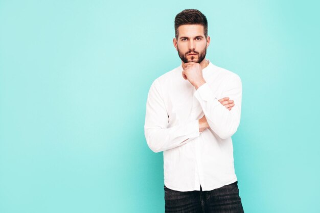 Retrato de guapo modelo confiado Hombre elegante sexy vestido con camisa blanca y jeans Hombre hipster de moda posando junto a la pared azul en el estudio Aislado