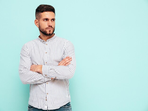 Retrato de guapo modelo confiado Hombre elegante sexy vestido con camisa y jeans Hombre hipster de moda posando junto a la pared azul en el estudio Brazos cruzados