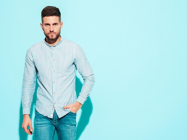 Retrato de guapo modelo confiado Hombre elegante sexy vestido con camisa y jeans Hombre hipster de moda posando junto a la pared azul en el estudio