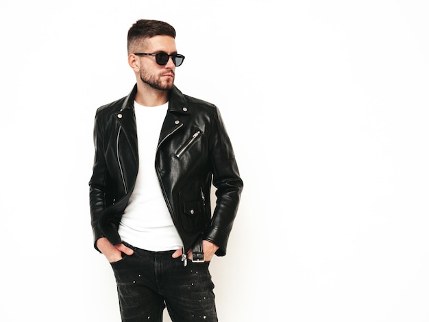 Retrato de guapo modelo confiado Hombre elegante sexy vestido con chaqueta de cuero de motociclista y jeans negros Hombre hipster de moda aislado en blanco en estudio En gafas de sol