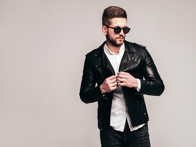 Retrato de guapo modelo confiado Hombre elegante sexy vestido con chaqueta de cuero de motociclista y jeans negros Hombre hipster de moda aislado sobre fondo gris en estudio con gafas de sol Aislado