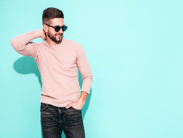 Retrato de guapo modelo confiado Hombre elegante sexy vestido con suéter rosa y jeans Hombre hipster de moda posando junto a la pared azul en el estudio En gafas de sol Aislado