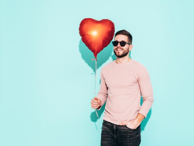 Retrato de guapo modelo confiado Hombre elegante sexy vestido con suéter rosa y jeans Hombre hipster de moda posando junto a la pared azul en el estudio En gafas de sol Sosteniendo globo de aire del corazón