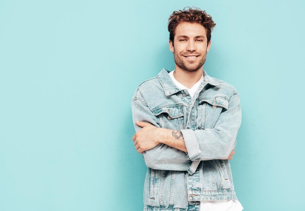 Retrato de guapo modelo lambersexual con estilo hipster con peinado rizado Hombre sexy vestido con chaqueta de jeans Hombre de moda aislado en la pared azul en el estudio