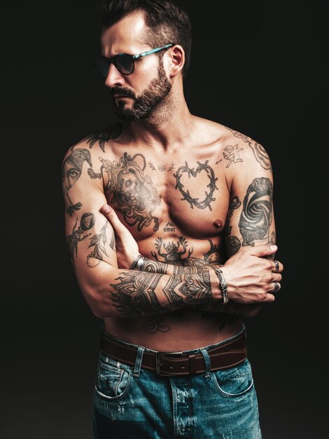 Retrato de guapo modelo lambersexual con estilo hipster seguro Hombre moderno sexy Torso desnudo con tatuajes Moda hombre posando en estudio sobre fondo oscuro en gafas de sol