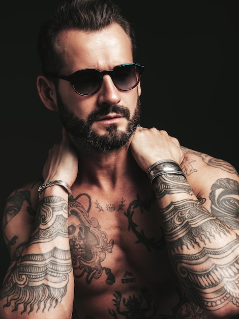 Retrato de guapo modelo lambersexual con estilo hipster seguro Hombre moderno sexy Torso desnudo con tatuajes Moda hombre posando en estudio sobre fondo oscuro en gafas de sol