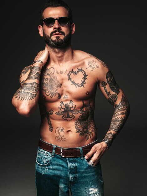 Retrato de guapo modelo lambersexual con estilo hipster seguro Hombre moderno sexy Torso desnudo con tatuajes Moda hombre posando en estudio sobre fondo oscuro en gafas de sol