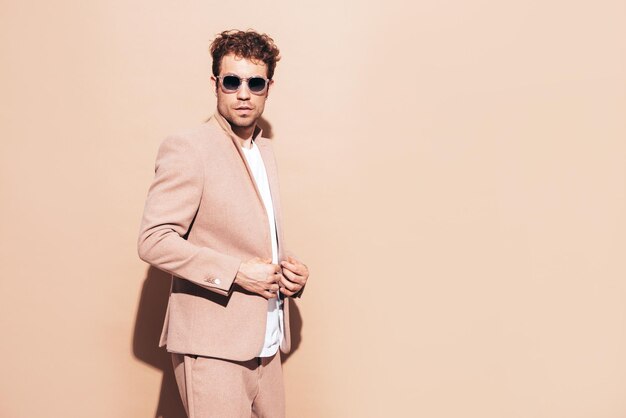 Retrato de guapo modelo lambersexual con estilo hipster seguro Hombre moderno sexy vestido con elegante traje beige Hombre de moda con peinado rizado posando en estudio Aislado en gafas de sol