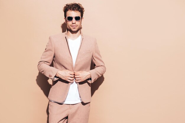 Retrato de guapo modelo lambersexual con estilo hipster seguro Hombre moderno sexy vestido con elegante traje beige Hombre de moda con peinado rizado posando en estudio Aislado en gafas de sol