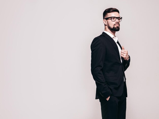 Retrato de guapo modelo lambersexual con estilo hipster seguro Hombre moderno sexy vestido con elegante traje negro Hombre de moda posando en el estudio cerca de la pared blanca Aislado en gris