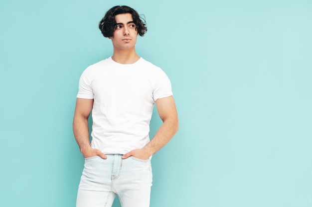 Foto gratuita retrato de guapo modelo lambersexual con estilo hipster seguro hombre sexy vestido con camiseta blanca de verano y ropa de jeans hombre de moda aislado en el estudio posando cerca de la pared azul