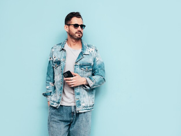 Retrato de guapo modelo lambersexual con estilo hipster seguro Hombre sexy vestido con chaqueta de jeans Hombre de moda aislado en la pared azul en el estudio En gafas de sol