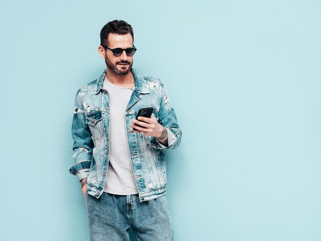 Retrato de guapo modelo lambersexual con estilo hipster seguro Hombre sexy vestido con chaqueta de jeans Hombre de moda aislado en la pared azul en el estudio En gafas de sol