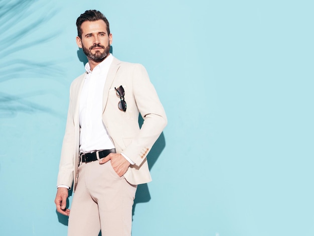 Foto gratuita retrato de guapo modelo lambersexual con estilo inconformista seguro hombre moderno sexy vestido con traje elegante blanco hombre de moda posando en el estudio cerca de la pared azul con gafas de sol