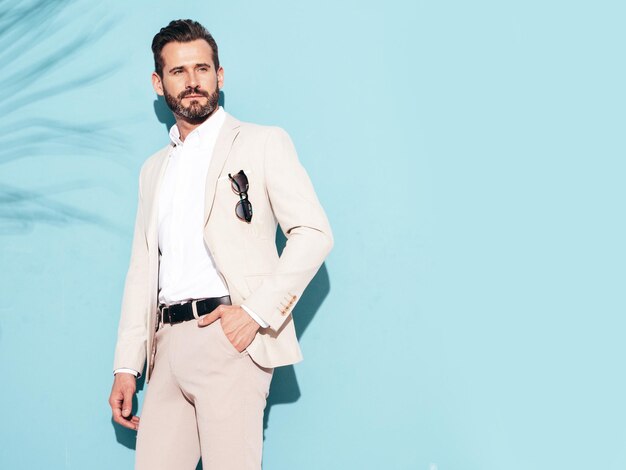 Retrato de guapo modelo lambersexual con estilo inconformista seguro Hombre moderno sexy vestido con traje elegante blanco Hombre de moda posando en el estudio cerca de la pared azul con gafas de sol